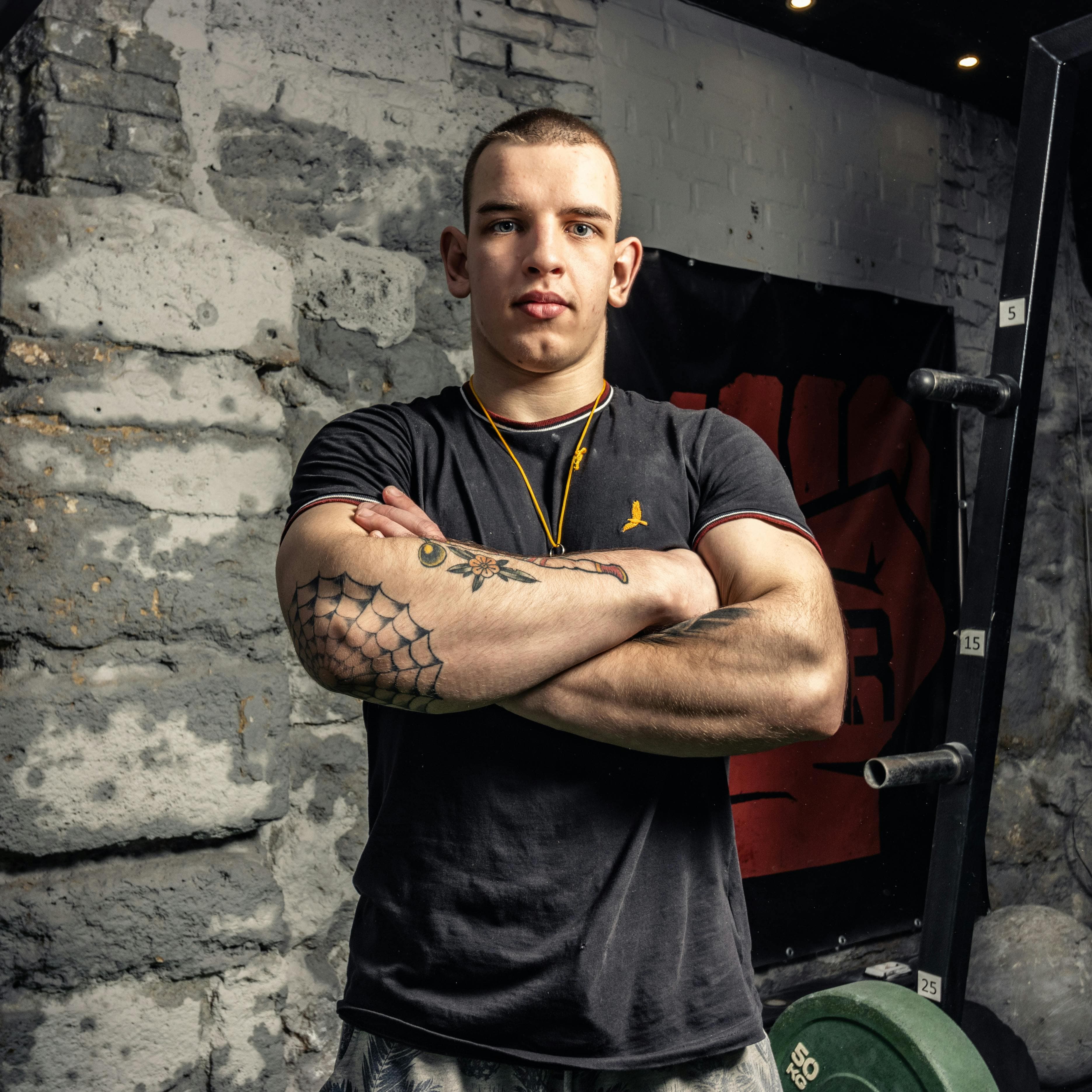 Strong man with tattoos standing in a gym, arms crossed confidently.