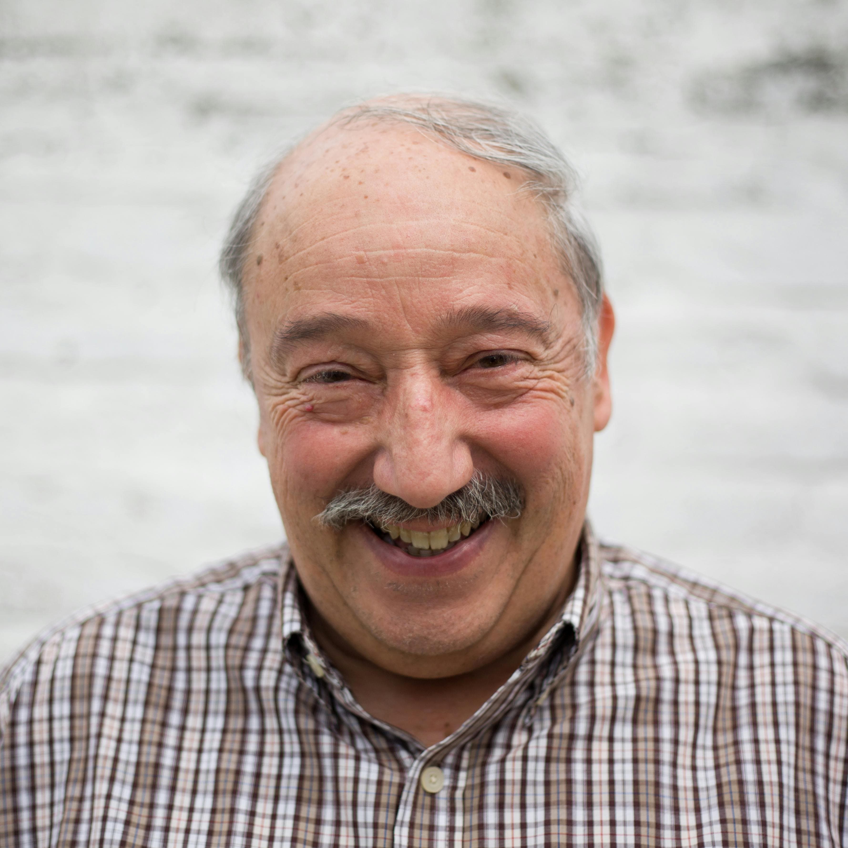 Elderly man with mustache smiling at the camera in an urban outdoor setting.