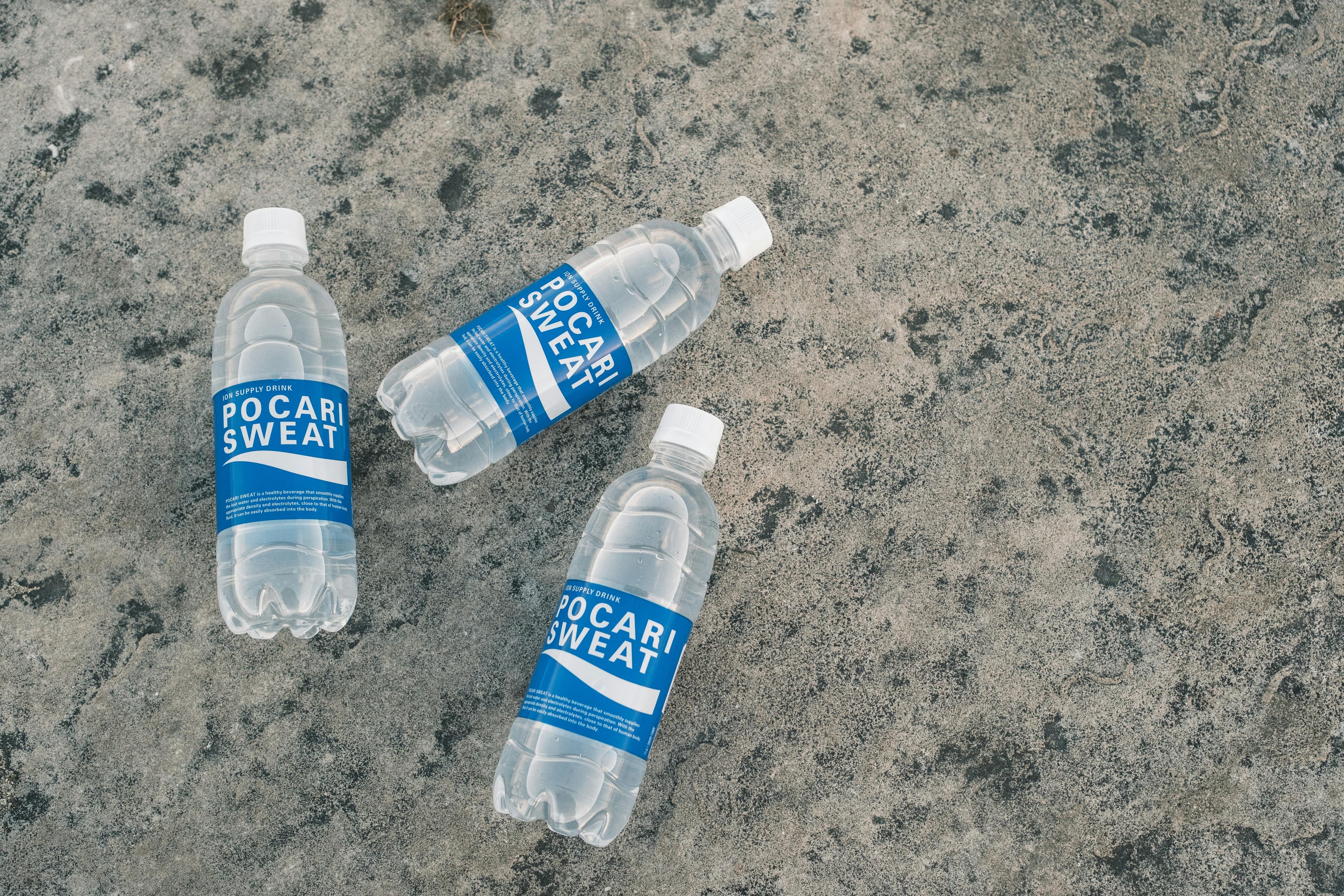 Tři láhve Pocari Sweat ležící na texturovaném skalnatém povrchu venku.