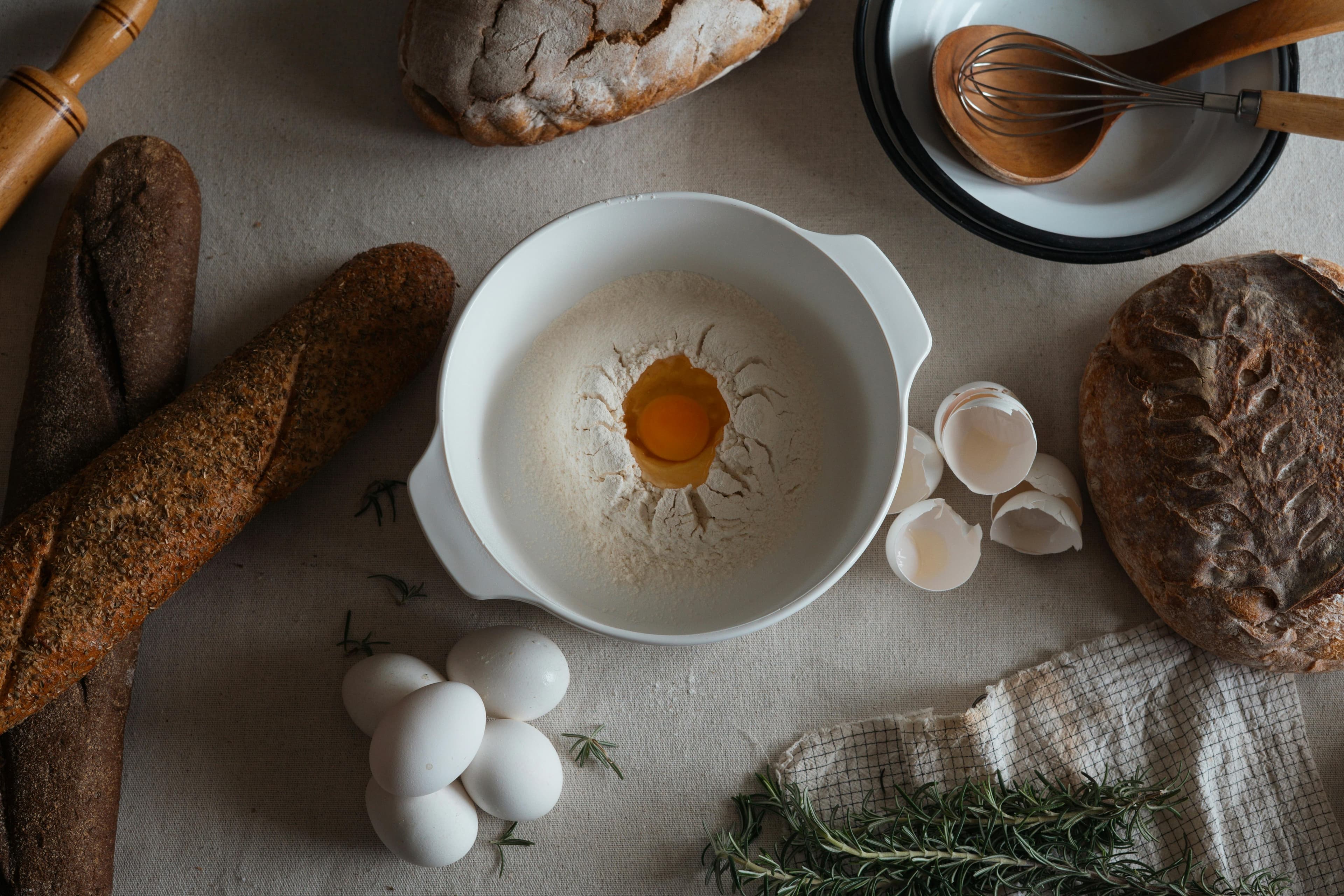 Camada plana de ingredientes para panificação, incluindo farinha, ovos, alecrim e pães variados.