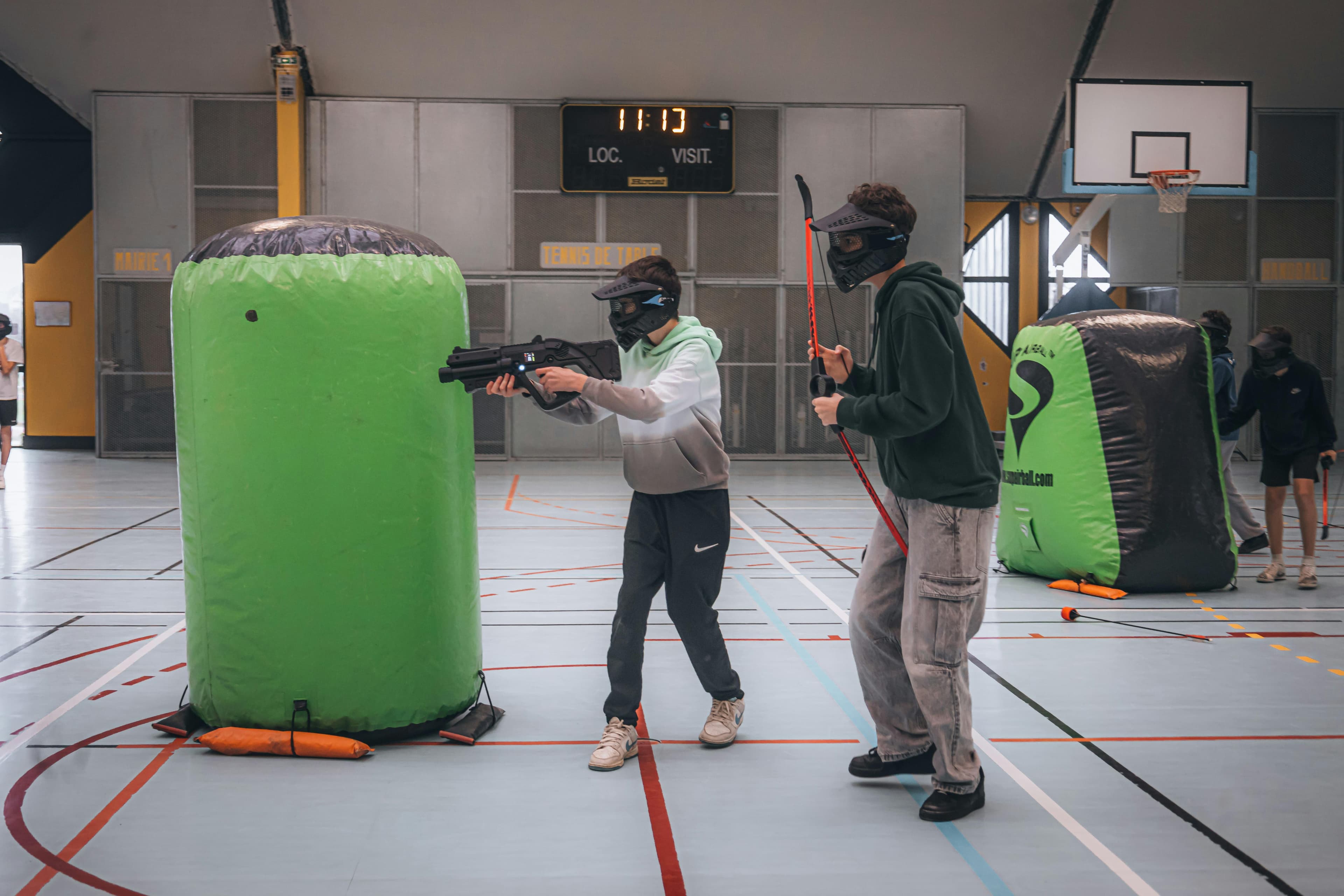 Dos personas jugando paintball en interiores usando máscaras y barreras inflables para cubrirse.