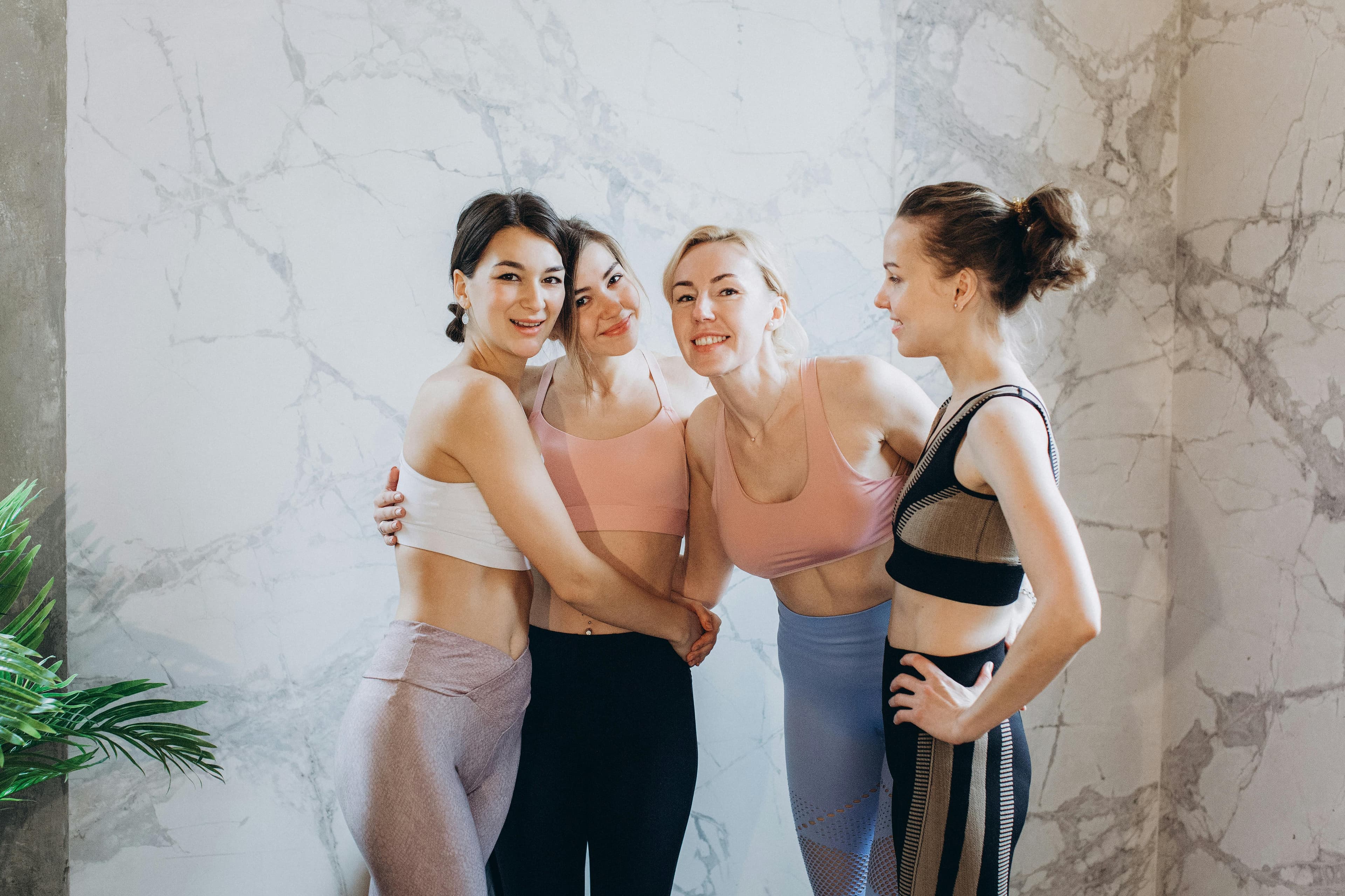 Four women in activewear smiling together, embodying friendship and fitness indoors.