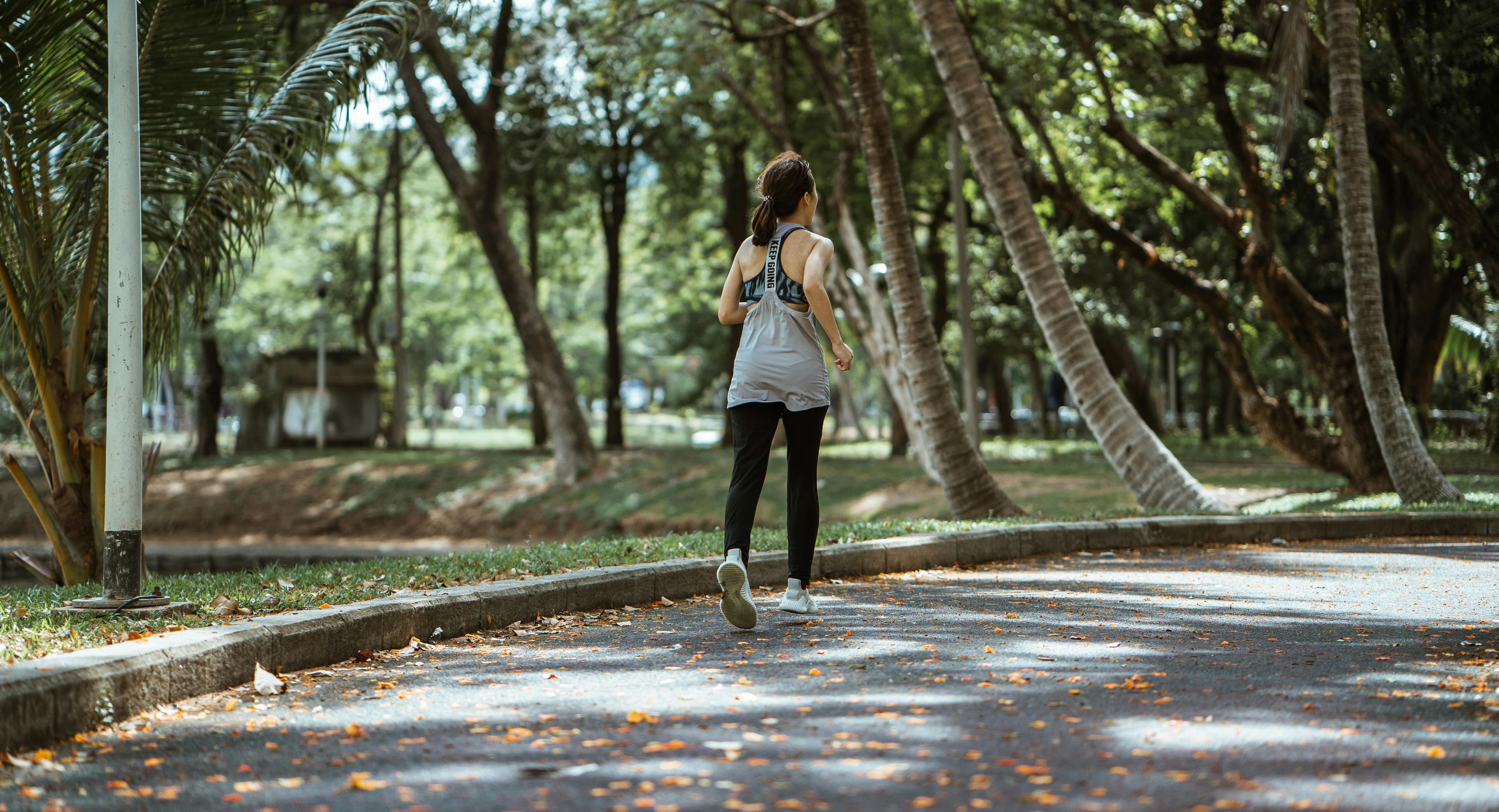 Back view of female in sportswear running on sidewalk in park in warm sunny day