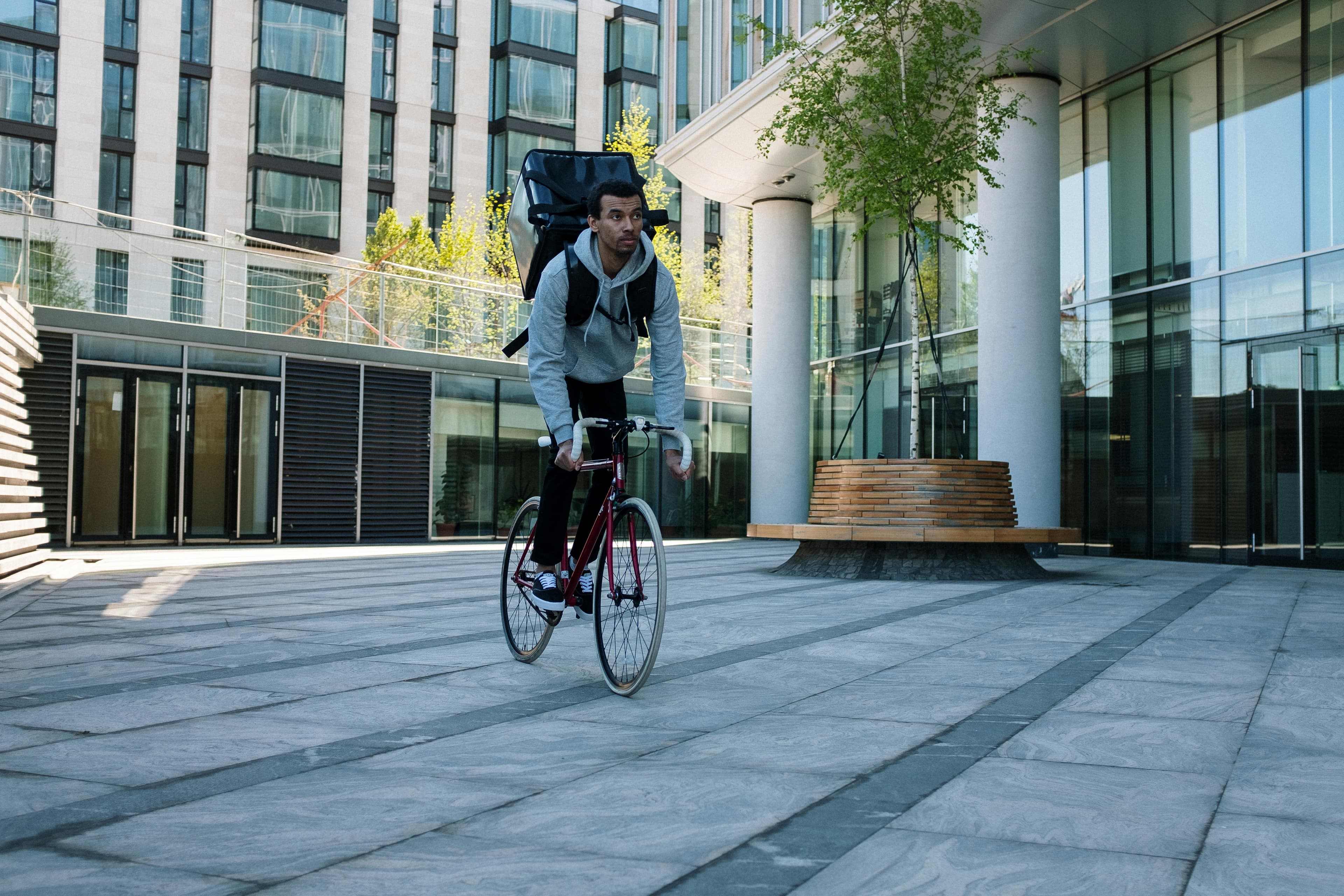 A cyclist with a backpack rides past modern architecture, epitomizing urban food delivery services.