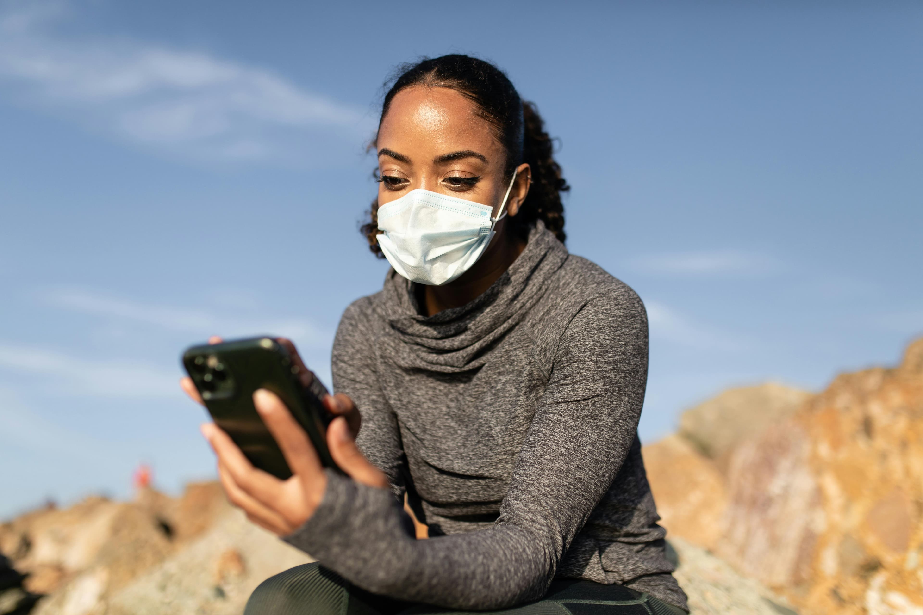 Woman wearing a mask uses smartphone outdoors, embracing new normal during pandemic.