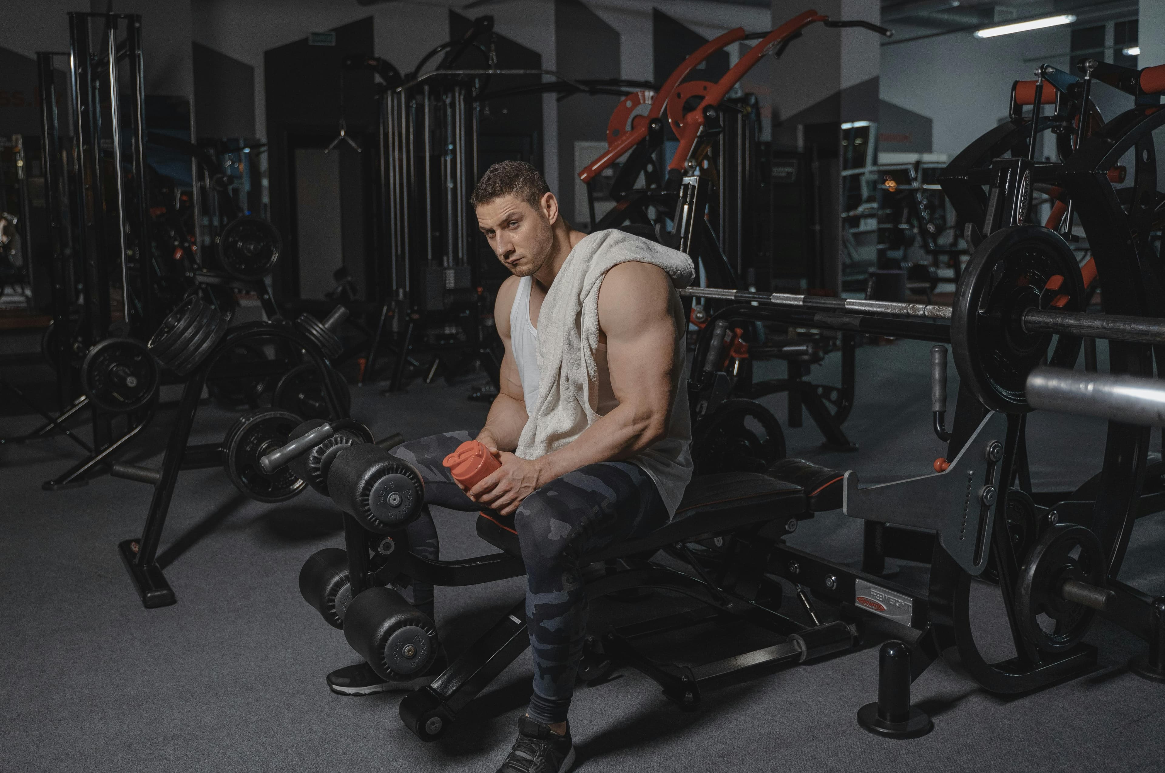 Man seated on bench in gym holding protein shaker, surrounded by equipment.