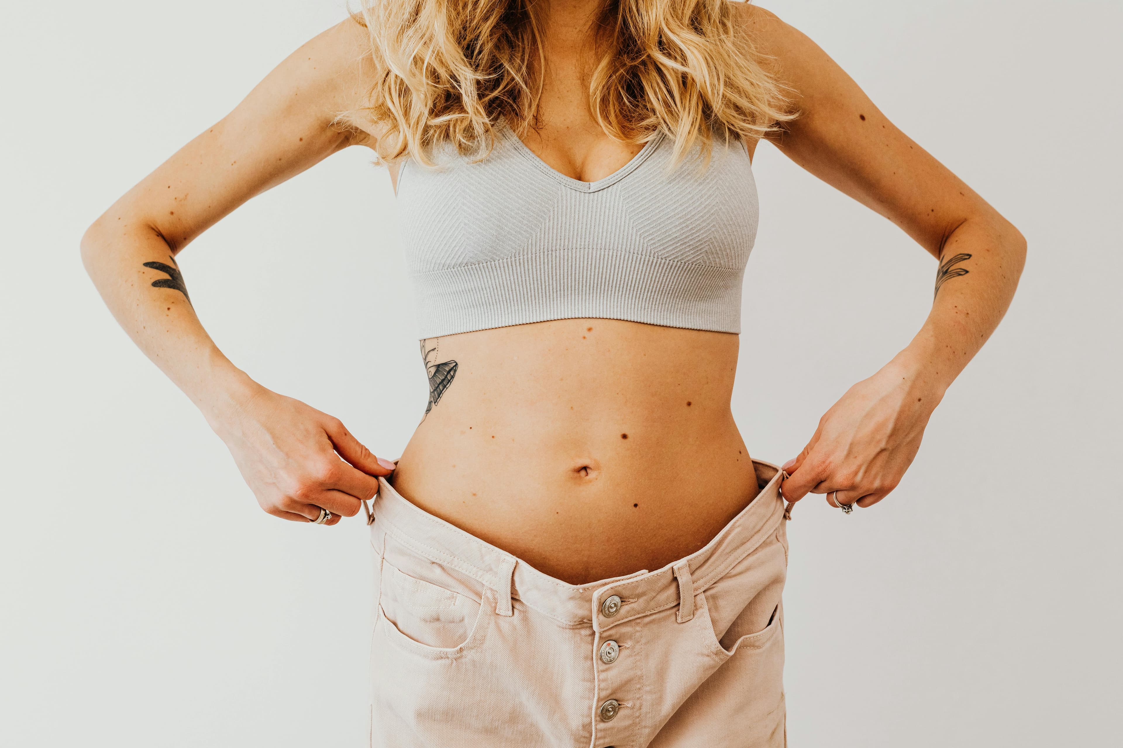 A woman displaying weight loss by wearing oversized pants and crop top, showing tattoos.