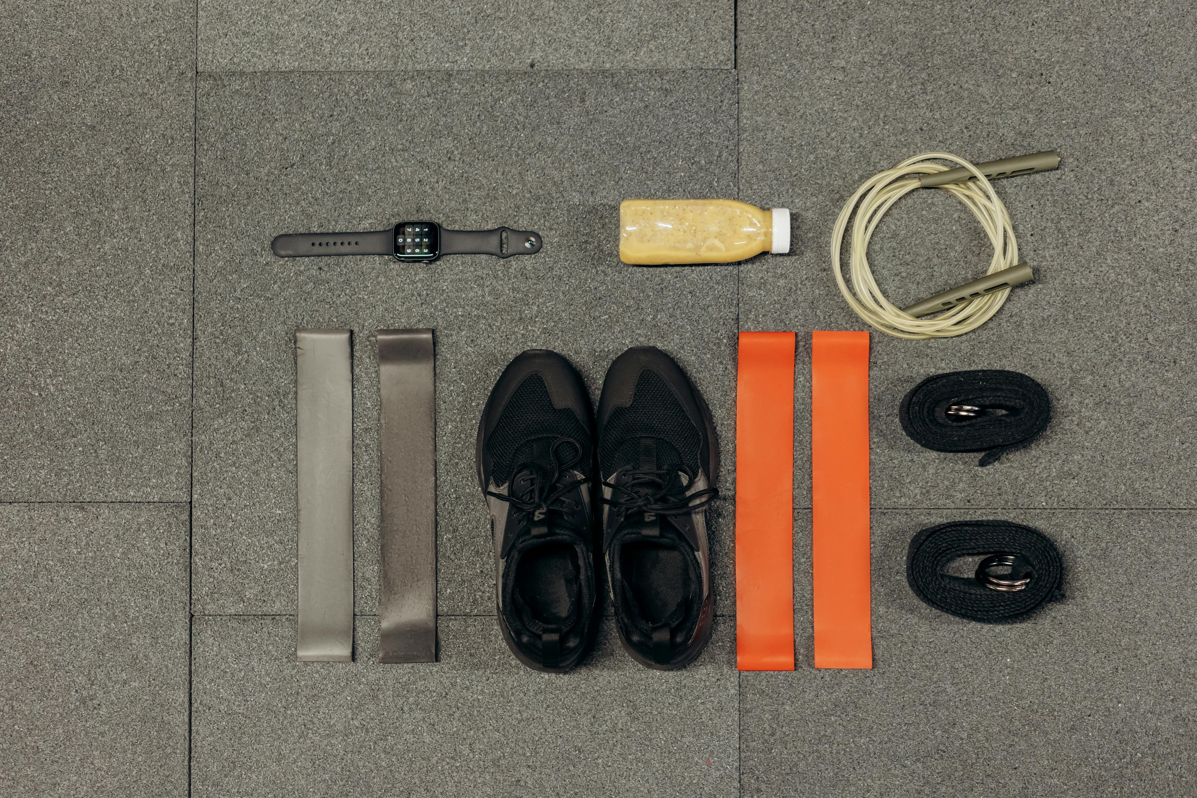 Flat lay of workout essentials including sneakers, bands, watch, rope, and healthy drink.