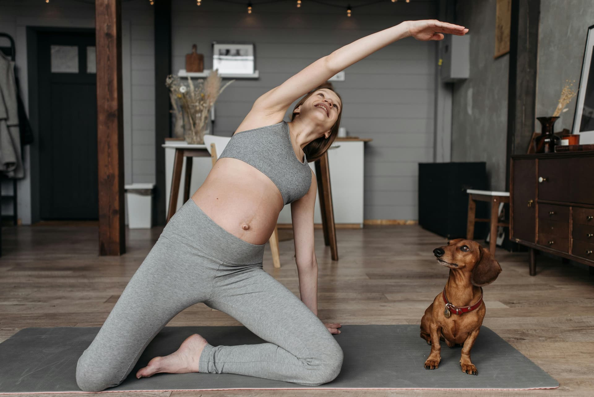 Pregnant woman practicing yoga with a dachshund indoors promoting healthy lifestyle.