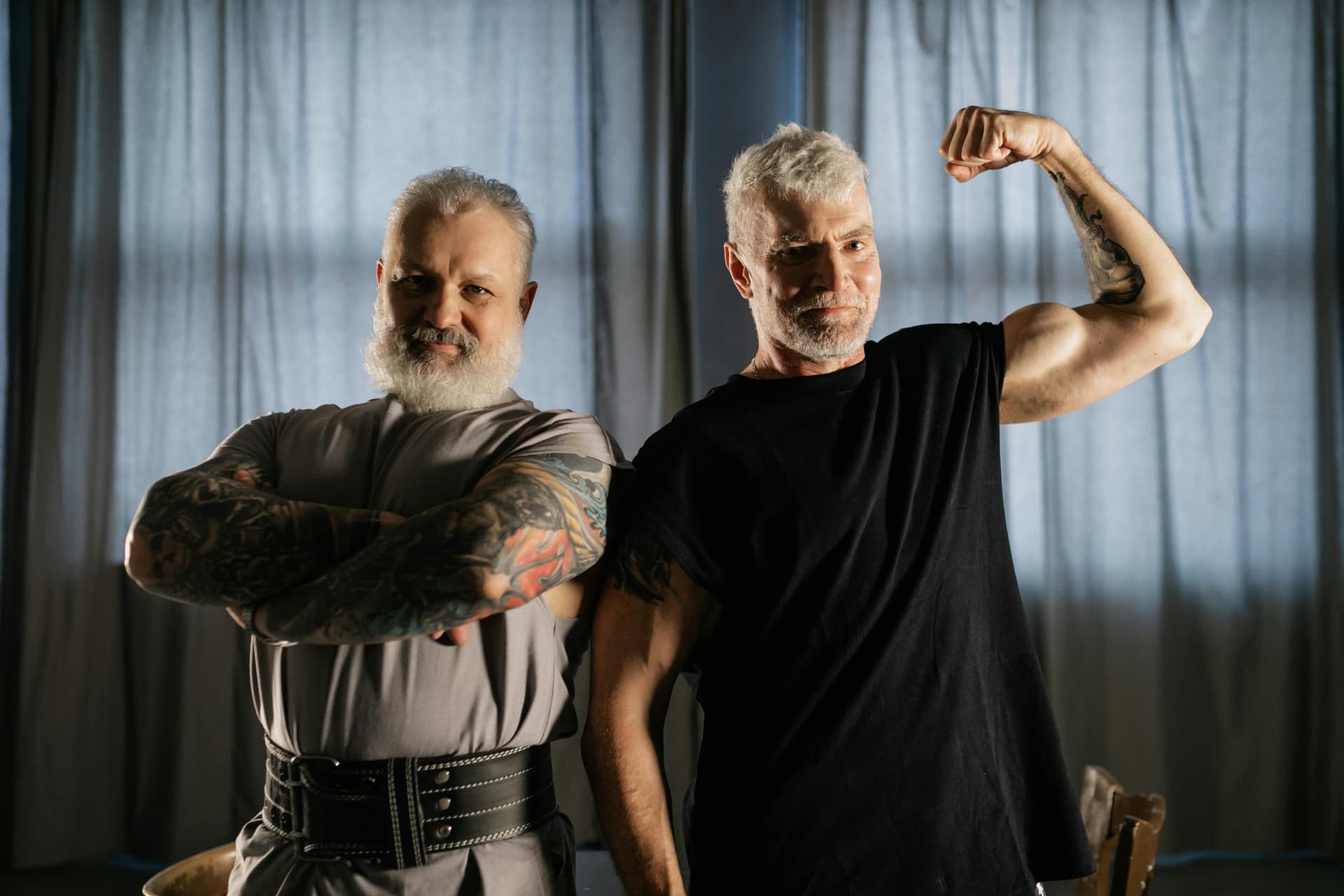 Two senior men confidently flexing and crossing arms indoors, showcasing strength and positive aging.