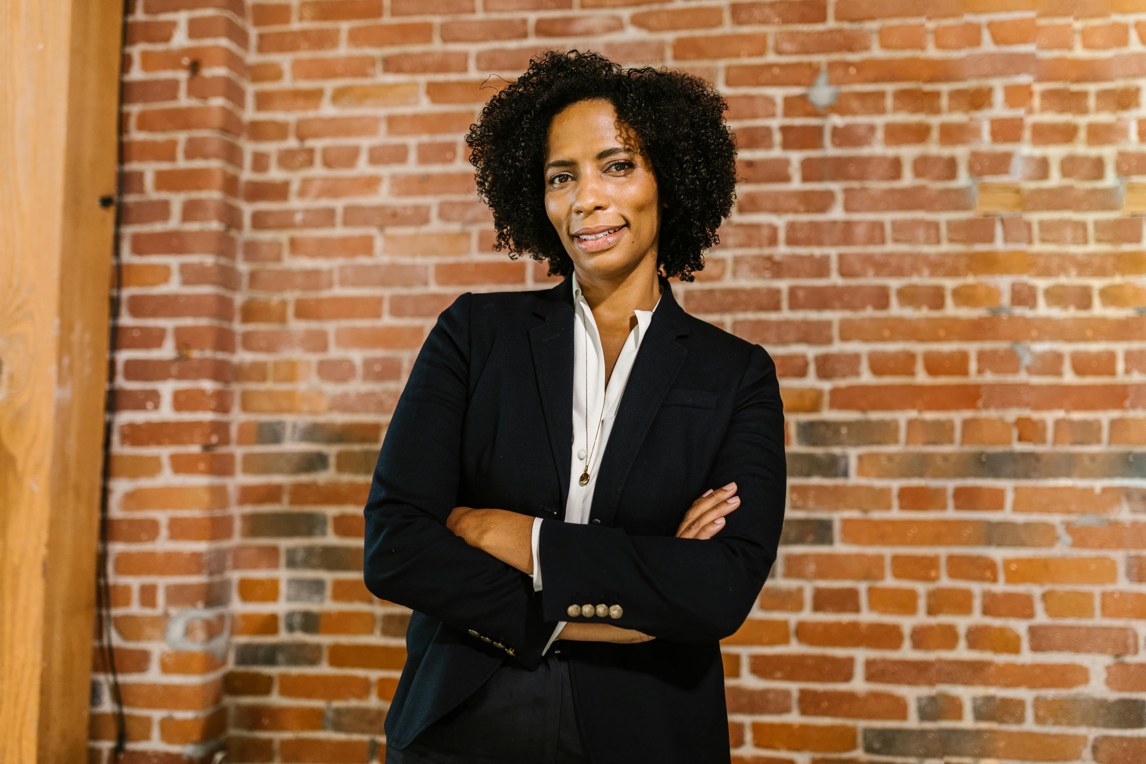 Professional black woman in corporate attire smiling confidently with arms crossed against a brick wall.