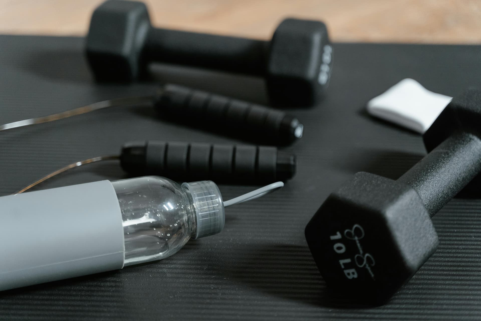 Close-up of gym essentials: dumbbells, jump rope, and water bottle on a mat.