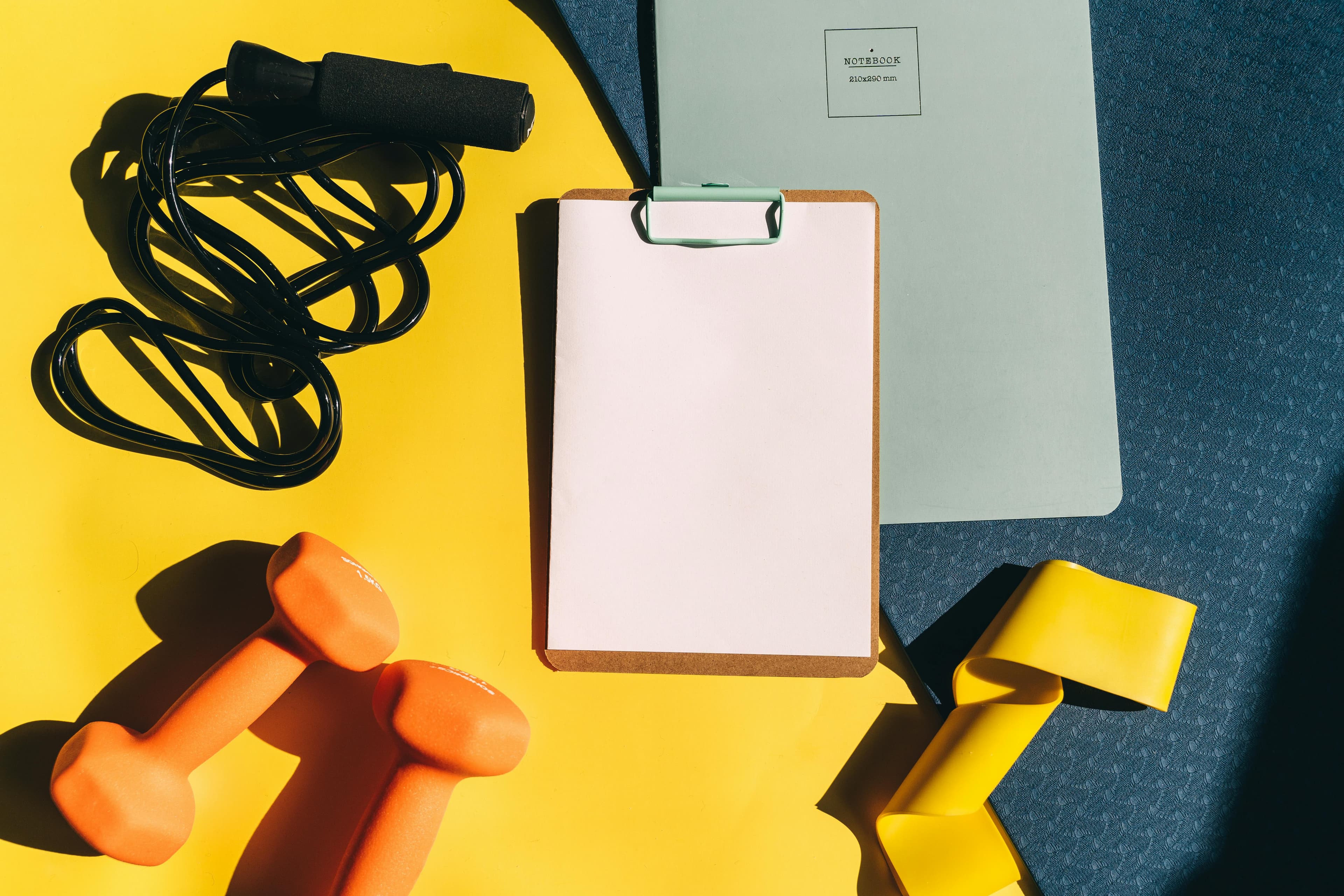 Flat lay of fitness equipment including dumbbells, jump rope, and clipboard on a yellow background.