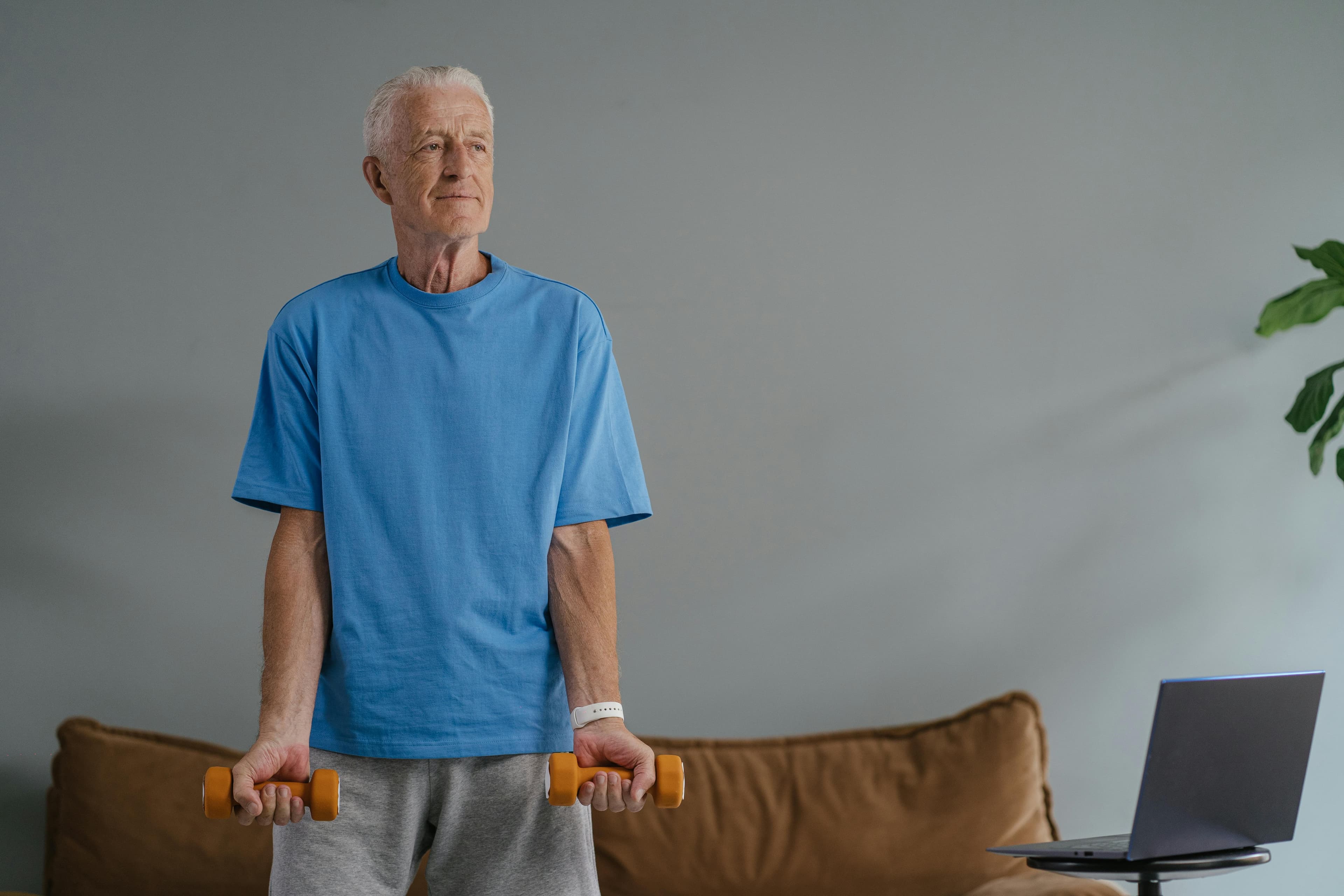 Elderly man in blue shirt lifting dumbbells near laptop, embracing home fitness.